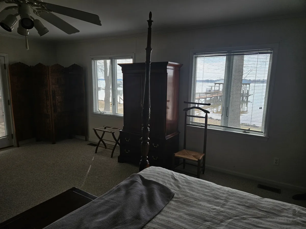 A beautifully appointed bedroom with a four-poster bed, large wooden wardrobe, and a window offering serene views of the water. The room’s calming color palette, decorative screen, and abundance of natural light create a peaceful and inviting atmosphere.