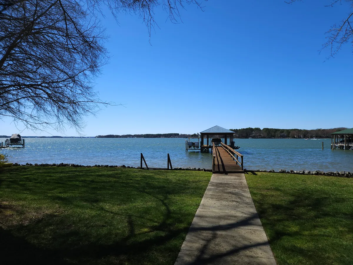 The image showcases a stunning, expansive view from the private dock, looking out over a calm, picturesque lake at sunset. The sky is painted with warm hues, and the tranquil water reflects the vibrant colors, creating a serene and inviting atmosphere. Lush green grass leads down to the dock, suggesting plenty of space for outdoor activities like barbecues, relaxing, or enjoying a morning coffee by the water.