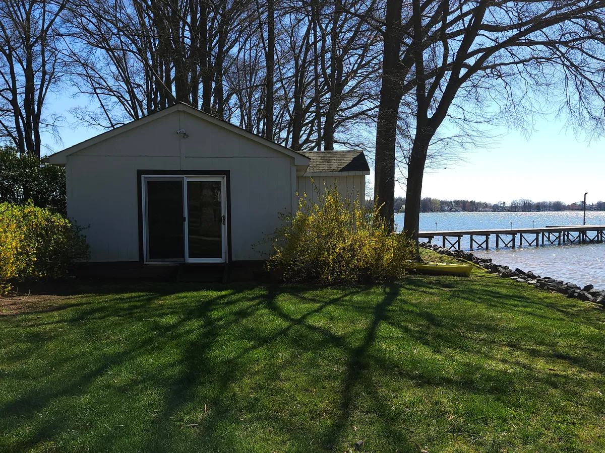 A small white building nestled near a yellow flowering bush, with the lake and a dock visible in the background. The setting exudes peacefulness and privacy, enhanced by lush greenery and a gentle waterfront ambiance ideal for quiet moments and evening reflections.