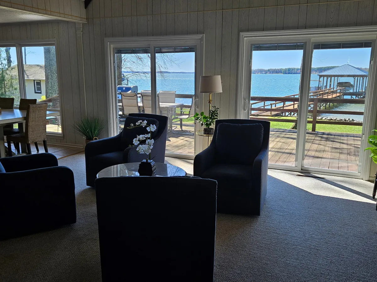 A bright, welcoming interior seating area with large windows framing the sparkling lake and deck outside. Plush chairs and a central coffee table create a comfortable space for guests to relax while staying visually connected to the lakefront and outdoor entertaining areas.