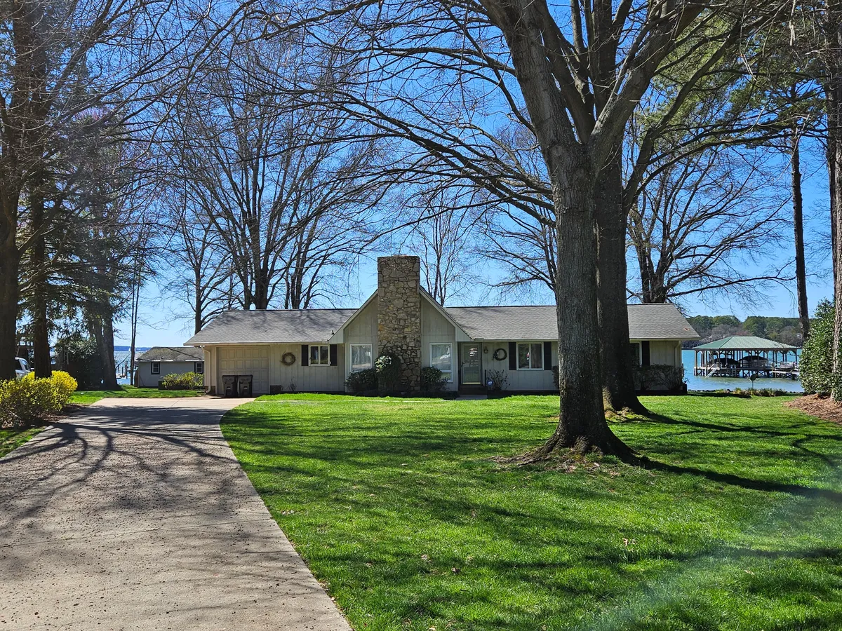 A welcoming exterior shot of the lakefront house, featuring a stone chimney, large trees, and a manicured lawn. The porch offers views across the water, and the scene conveys a space perfect for family gatherings, outdoor barbecues, and relaxing after a day of adventure.