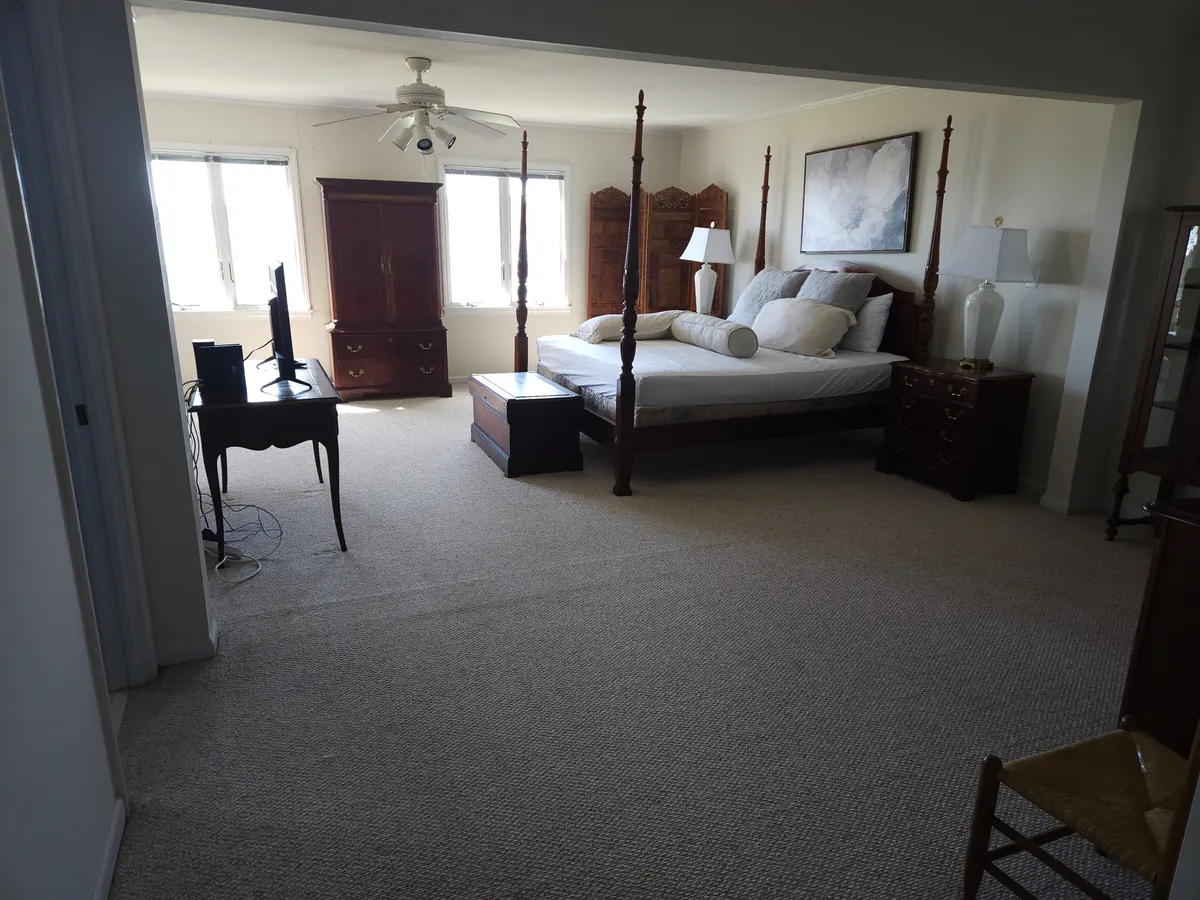 A spacious bedroom featuring a four-poster bed, large windows that flood the room with natural light, stylish nightstands, a dresser, and a television. The space feels cozy and peaceful, ideal for rest and quiet moments.
