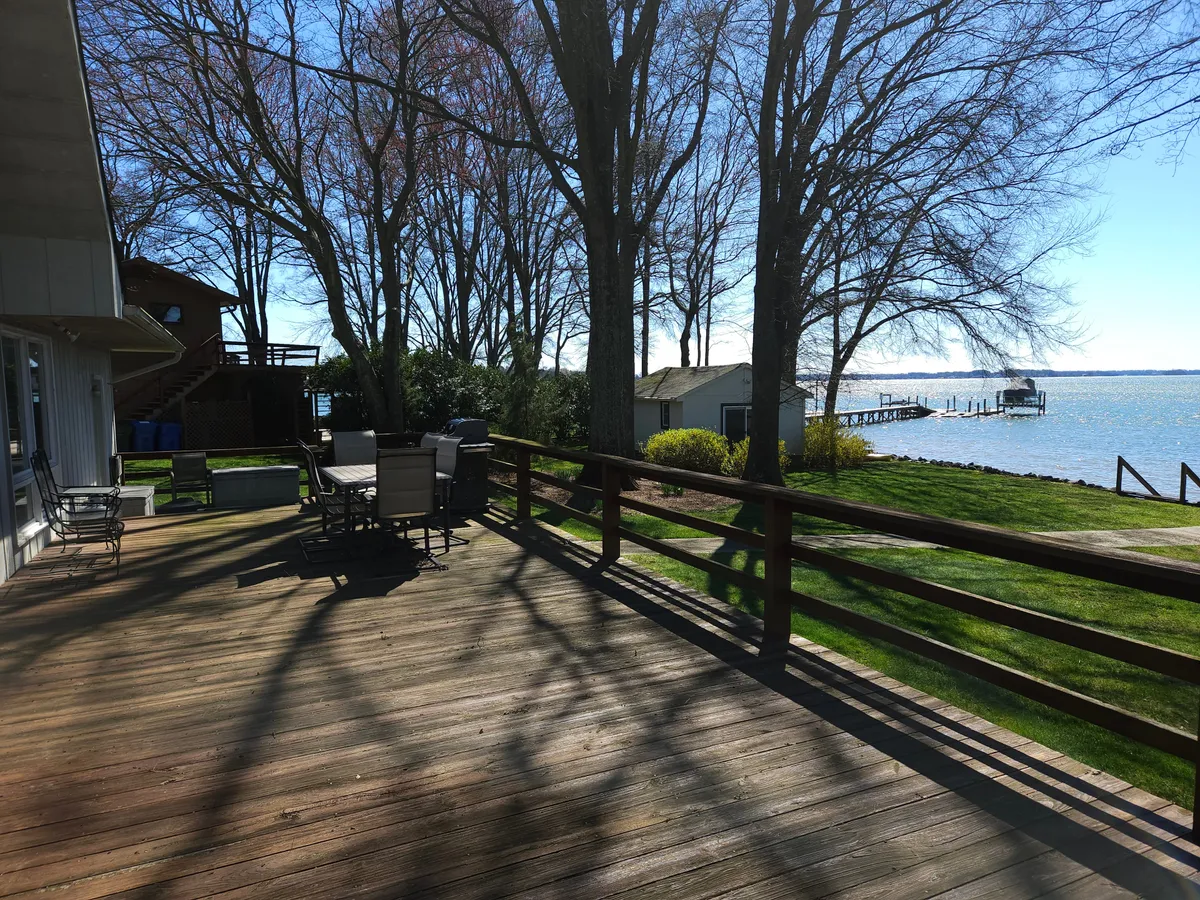 A spacious wooden deck featuring a stylish outdoor dining set under the shade of tall trees, overlooking the serene lake. The deck offers ample space for gatherings, with a view that seamlessly merges the comforts of home with nature. The water glimmers in the background, setting a perfect scene for dining and entertaining.